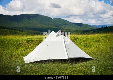 Tente touristique blanche au soleil, au sommet d'une colline herbeuse, entourée de jeunes pins. Vue panoramique sur les montagnes luxuriantes et vallonnées sous un ciel bleu vif avec des nuages dispersés, créant un endroit de camping idyllique. Banque D'Images