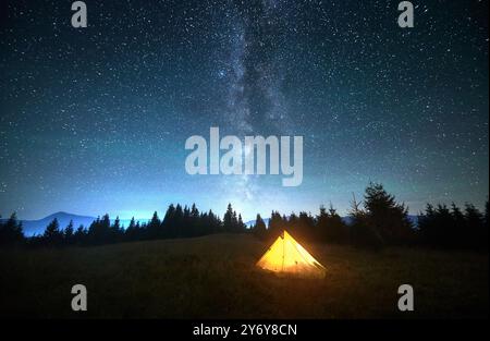 Tente touristique jaune incandescente sur une colline herbeuse sous un ciel étoilé époustouflant avec la voie lactée bien visible. Silhouette de pins et de montagnes. Beauté sereine de la nuit claire dans la nature. Banque D'Images