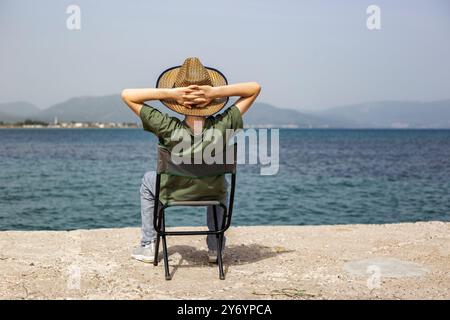 Vue arrière d'une personne assise avec les bras croisés sur la tête au bord de la mer Banque D'Images