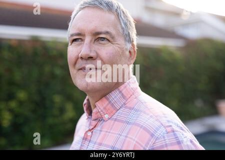 Homme senior souriant en chemise à carreaux appréciant le temps en plein air dans le jardin, à la maison Banque D'Images