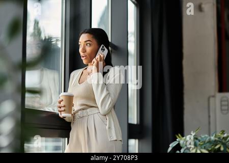 Une jeune belle femme se tient près d'une grande fenêtre, engagée dans une conversation téléphonique animée. Banque D'Images