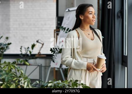 Une jeune femme se tient dans un bureau, réfléchissant à sa journée avec une tasse à café à la main. Banque D'Images