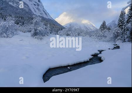 Le filtre de lumière douce à travers les nuages illumine un paysage enneigé avec un ruisseau qui coule doucement au milieu de montagnes imposantes. Banque D'Images