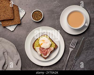 Photographie alimentaire du petit déjeuner nutritionnel, pain de seigle au levain avec de la purée d'avocat, prosciutto, bacon, oeuf poché bouilli, thé avec du lait Banque D'Images