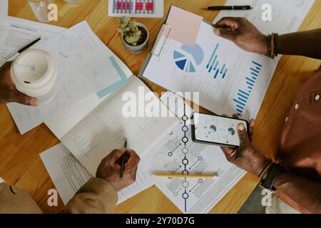 Top down shot de mains masculines méconnaissables avec smartphone et tasse thermique de café, table en bois avec des papiers avec des diagrammes et des schémas Banque D'Images