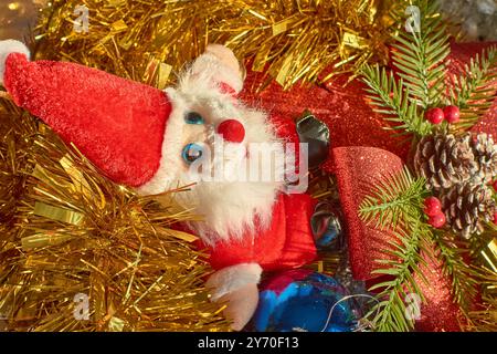 Jouet en peluche du Père Noël assis au milieu d'une scène de Noël festive. La figure du Père Noël, avec sa barbe blanche moelleuse, son costume rouge vif et ses yeux bleus, est surr Banque D'Images