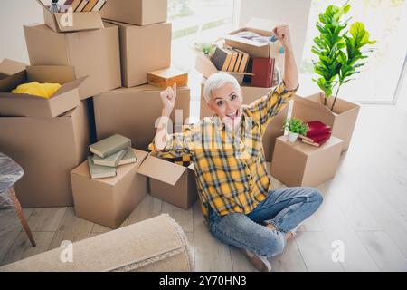 Photo de vue en grand angle de vue de belle femme âgée soulever poings assis sol porter chemise à carreaux déplacer déménager nouvel appartement à l'intérieur Banque D'Images