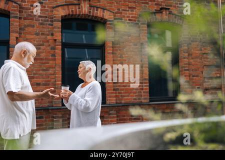 Vue latérale d'un couple âgé discutant joyeusement dans la rue urbaine avec des tasses à café, savourant la chaude journée d'été. Bel homme senior et femme souriant Banque D'Images