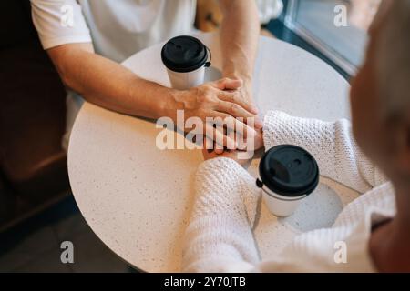 Gros plan sur le dessus d'un couple d'aînés heureux méconnaissable partageant une conversation chaleureuse autour d'un café, tenant les mains entrelacées, rayonnant d'amour Banque D'Images
