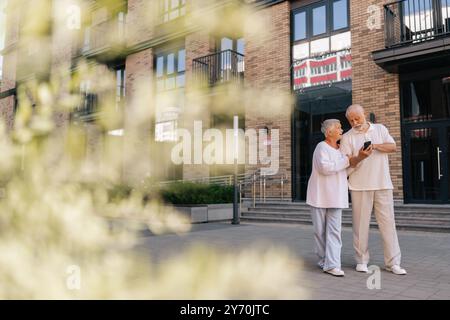 Grand plan à distance d'un beau couple de personnes âgées explorant de nouvelles rues de la ville, en comptant sur le smartphone pour les directions. Homme et femme âgés aux cheveux gris Banque D'Images