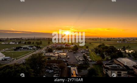 Alors que le soleil se couche au-delà de l'horizon, des teintes orange et jaune vibrantes remplissent le ciel, projetant une lumière chaude sur les champs et un parking occupé, créant une atmosphère sereine en soirée dans la campagne. Banque D'Images