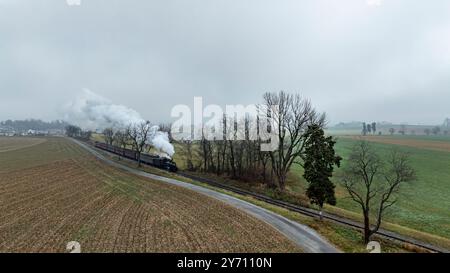 Un train à vapeur d'époque glisse le long de voies ferrées bordées de champs et d'arbres sous un ciel nuageux et chargé de brouillard. Le paysage met en valeur un environnement rural serein dans la lumière tôt le matin. Banque D'Images