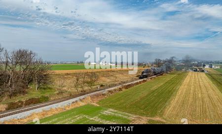 Une locomotive à vapeur classique souffle de la fumée bleu-gris alors qu'elle se déplace le long des voies ferrées, entourée de champs verdoyants et de terres agricoles. Le ciel dégagé améliore le paysage rural tranquille. Banque D'Images