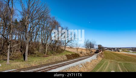 Une locomotive à vapeur d'époque fait son chemin le long de la voie ferrée, entourée d'arbres et de collines ondulantes. Le ciel est dégagé et offre une vue imprenable sur le paysage. Banque D'Images