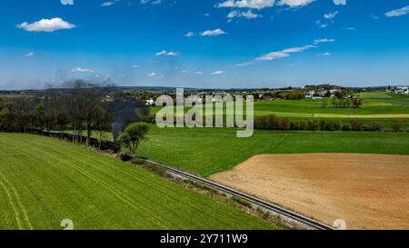 Une locomotive à vapeur souffle des nuages de fumée noire tout en voyageant à travers des champs verts vibrants sous un ciel bleu vif. Le paysage rural paisible présente des collines lointaines et des terres agricoles. Banque D'Images