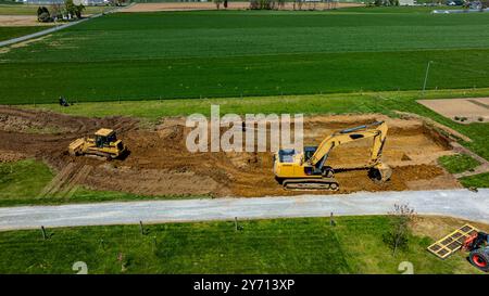 De la machinerie lourde, dont une excavatrice et un bulldozer, fonctionne sur un chantier de construction dans une zone rurale verte. Le sol est déplacé par temps clair, mettant en valeur le site actif. Banque D'Images