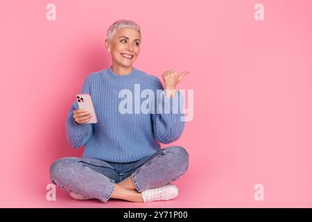 Photo pleine longueur du point de regard de dame retraitée espace vide assis dispositif de maintien de plancher porter une tenue de tricot bleu isolé sur fond de couleur rose Banque D'Images