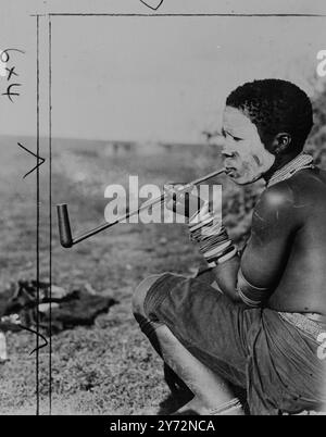 LA LÉGENDE ORIGINALE de 1947 SUIT : étude dans le nez au Parlement de couverture rouge. Fumer le reste dans la pipe de paix. Visage enduit d'argile (un signe traditionnel de deuil), sa longue pipe en bois d'olivier à tige dessinant gentiment, une femme de la tribu Xhosa South African Transkeian natives squat et pense à son cher disparu. Normalement, elle porte une épaisse couverture rouge ocre, l'insigne de la tribu. Le roi et la reine de Grande-Bretagne ont rencontré quelques-uns des menfolk des tribus, des représentants du célèbre Parlement natif de Transkeian, lors de leur visite à Umtata, capitale de la réserve indigène Red Blanket. 4 mars 1947 Banque D'Images