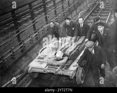 Les pompiers, la police et les cheminots ont utilisé des appareils de coupe spéciaux pour extirper pour L.P.T.B. des employés soupçonnés d'avoir été piégés dans plusieurs voitures du train de métro qui a colmaté avec un autre train dans la section souterraine de la section est de la ligne centrale qui a été ouverte au public hier. Les deux trains étaient vides de passagers au moment de l'accident qui s'est produit sur un tronçon de ligne en boucle qui est utilisé pour faire tourner les trains au-delà de la gare réelle de Stratford. Images : un des hommes blessés après avoir été libéré de l'épave, étant sorti du Banque D'Images