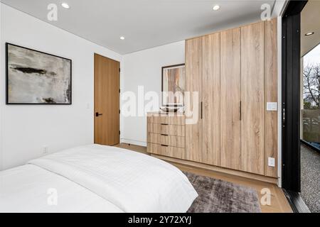 L'intérieur moderne de la chambre comprend une armoire en bois, des œuvres d'art encadrées et un lit confortable avec des draps blancs, bénéficiant d'une lumière naturelle provenant d'une grande fenêtre. Banque D'Images