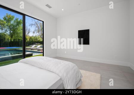 Chambre moderne minimaliste avec une grande fenêtre donnant sur la piscine et un coin salon extérieur, avec murs blancs et décoration simple. Banque D'Images