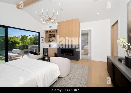 Chambre moderne avec grandes fenêtres, balcon avec vue sur la verdure et décor contemporain comprenant lustre, cheminée et touches de bois. Banque D'Images