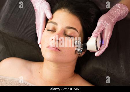 Une esthéticienne utilise un dispositif à micro-courant sur la joue d'un client, effectuant un massage facial pour améliorer la texture de la peau. Banque D'Images