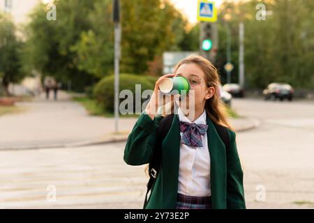 Une jeune fille dans un blazer vert buvant dans une tasse verte tout en marchant à travers une rue, avec un passage pour piétons et des feux de circulation en arrière-plan. Banque D'Images