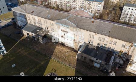 Une vue aérienne d'un bâtiment délabré entouré d'herbe envahie et de débris. A proximité, il y a des bâtiments résidentiels en arrière-plan, contraste Banque D'Images