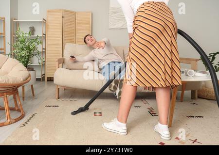 Mari paresseux regardant la télévision pendant que sa femme aspirateur la moquette à la maison Banque D'Images