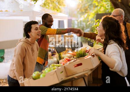 Vendeur féminin joyeux offrant des fruits et légumes fraîchement récoltés au consommateur caucasien au stand de la foire de la ferme de récolte. Humeur positive au marché des agriculteurs, clients multiraciaux achetant des produits biologiques. Banque D'Images