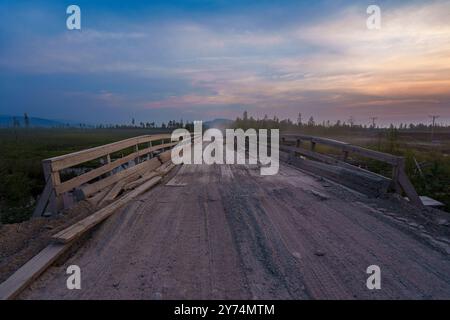 Un pont en bois altéré avec des garde-corps endommagés s'étend à travers un paysage rural au crépuscule. La scène est paisible et calme, avec un ciel doux du soir i Banque D'Images