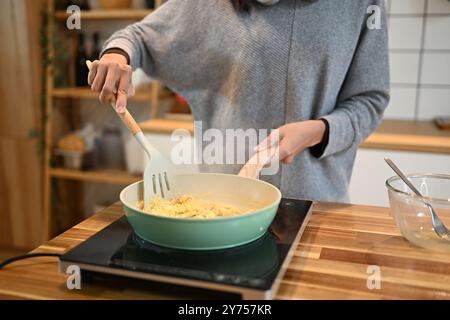 Jeune femme coupée cuisinant des œufs brouillés dans une poêle verte sur le comptoir de la cuisine Banque D'Images