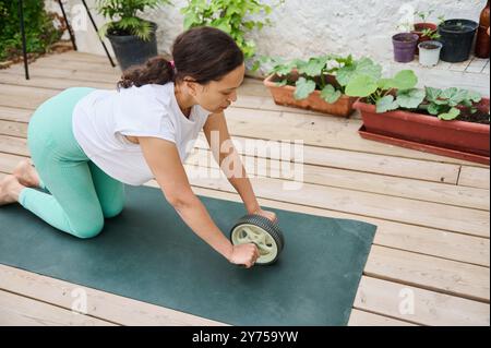 Femme concentrée utilisant un rouleau ab sur un tapis de yoga dans un cadre extérieur serein avec de la verdure, favorisant un mode de vie sain et actif. Banque D'Images