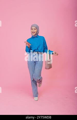 Une jeune femme élégante dans une chemise bleue et un Jean pose gracieusement sur un fond rose vif Banque D'Images