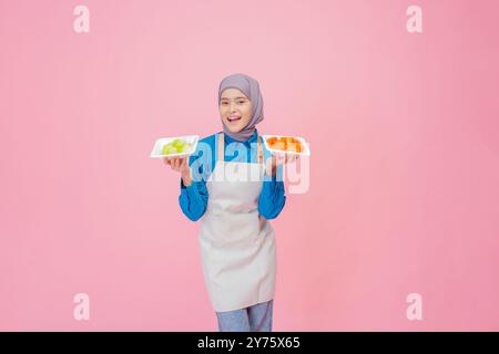Une femme joyeuse dans un tablier tient en toute confiance des assiettes de légumes frais sur un fond rose Banque D'Images