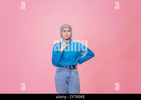 Une femme réfléchie vêtue d'une chemise bleue et d'un pantalon en denim pose gracieusement sur un fond rose Banque D'Images