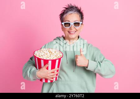 Photo de confiant attirant Lady Wear kaki sweat à capuche montrant pouce vers le haut regarder le film manger pop corn isolé fond de couleur rose Banque D'Images