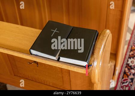 Livre de prières noir avec un symbole de croix chrétienne sur elle reposant sur un banc de banc d'église, plan détaillé en gros plan, personne. Bible, hymne, évangile, petite prière b Banque D'Images