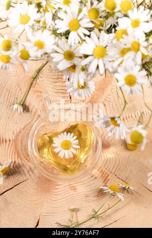 Tisane de camomille en forme de coeur dans une tasse en verre avec des fleurs sur fond de souche en bois. Vue de dessus. La boisson à base de plantes est bonne pour le sommeil, la peau, les cheveux c Banque D'Images