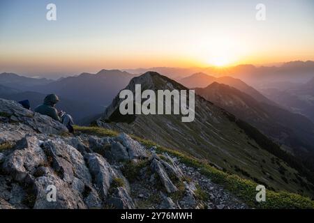 Un randonneur est assis au sommet du mont Hinteres Sonnwendjoch et regarde le lever du soleil sur les montagnes Kaiser, Tyrol, Autriche Banque D'Images