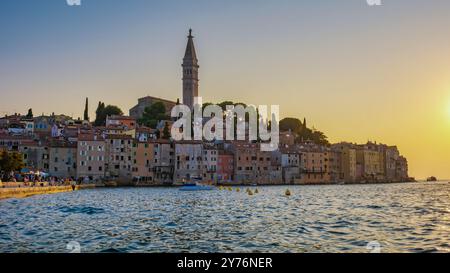 Alors que le soleil plonge sous l'horizon, le front de mer de Rovinj Croatie prend vie avec des teintes chaudes, mettant en valeur des bâtiments colorés, une église imposante et des eaux tranquilles invitant à l'exploration le long de la côte sereine. Banque D'Images