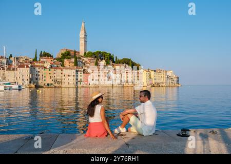 Un couple profite d'un moment paisible au bord de l'eau à Rovinj, en Croatie, avec des bâtiments colorés reflétant dans la mer calme et le clocher emblématique debout contre le ciel bleu clair. Banque D'Images