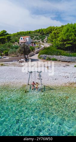 Deux amis se détendent sur une balançoire par les belles eaux turquoises de l'île de Brac, en Croatie, entouré d'une végétation luxuriante et d'une charmante maison côtière, créant une ambiance d'escapade estivale parfaite. Banque D'Images