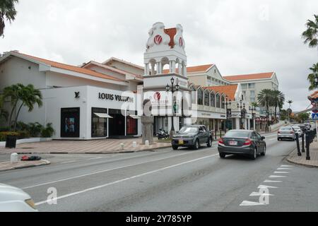 Route principale avec des bâtiments modernes avec des boutiques de luxe, des palmiers et des voitures neuves à Oranjestad à Aruba au printemps sous ciel couvert. Banque D'Images