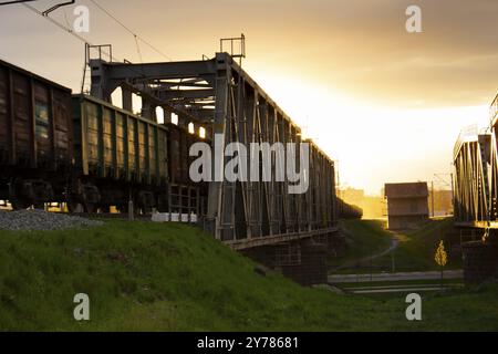 Mise au point sélective.Wagons de marchandises sur un pont ferroviaire tirés par un train.Lumière du soleil du soir et reflet de la lumière orange du soir de la structure métallique Banque D'Images