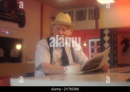 Homme senior portant un chapeau de paille et vérifiant le journal au comptoir du café, immergé dans un cadre de café confortable avec un éclairage doux et des couleurs chaudes, créant une atmosphère accueillante idéale pour la détente et le plaisir Banque D'Images