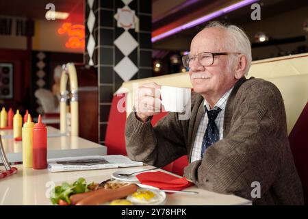 Homme âgé savourant une tasse de café assis dans le restaurant rétro, mettant en valeur une ambiance nostalgique avec un décor classique, entouré d'articles pour le petit déjeuner et de matériel de lecture Banque D'Images