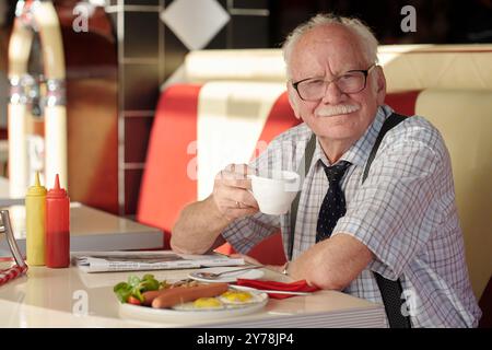 Portrait d'un homme âgé avec moustache assis dans le restaurant, souriant tout en tenant une tasse à café, avec de la nourriture sur la table et rouge, cabine blanche en arrière-plan Banque D'Images