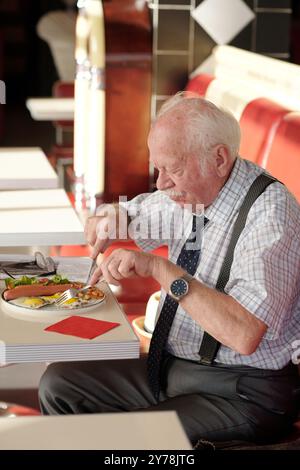 Homme âgé appréciant le repas tout en étant assis dans le cadre confortable du restaurant. Vêtu d'une tenue habillée et utilisant des ustensiles, il se concentre sur sa nourriture sans distractions Banque D'Images